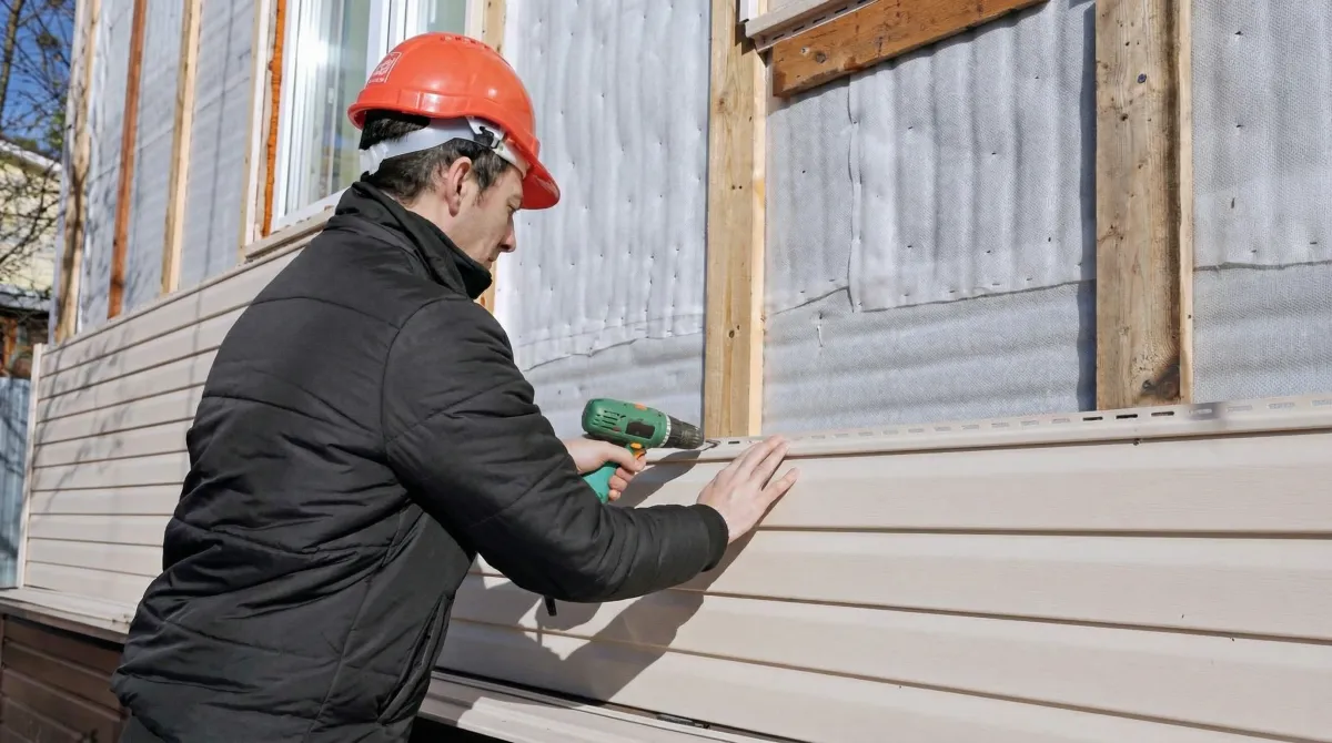 Siding replacement professional using a power drill to install new tan vinyl siding on a home exterior in Tuscaloosa, AL